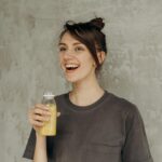 Young woman smiling while holding a refreshing bottle of orange juice indoors.